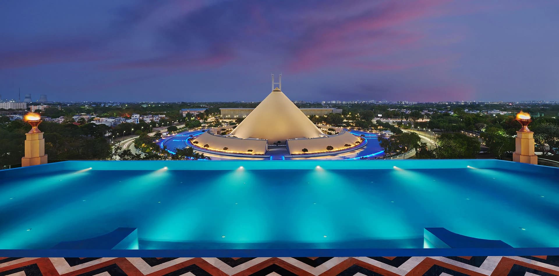 The Leela Gandhinagar rooftop infinity pool at twilight, overlooking the illuminated Dandi Kutir cone